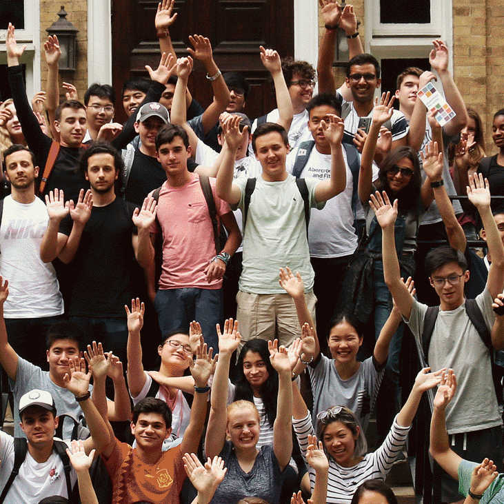 Large group of students in two rows, standing and sitting with their arms raised up, smiling in front of Cumberland House.