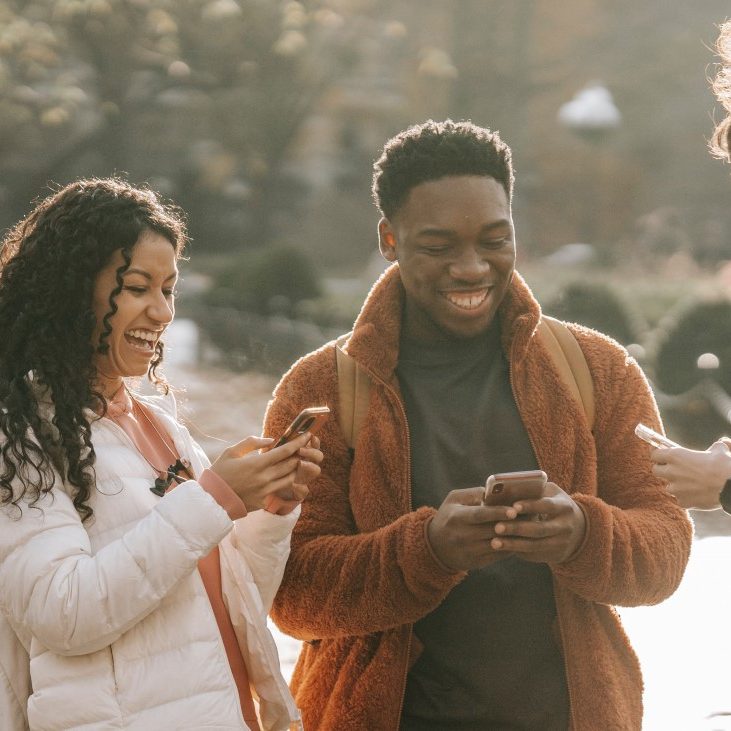 Three students standing outside together, looking at their phones and smiling.