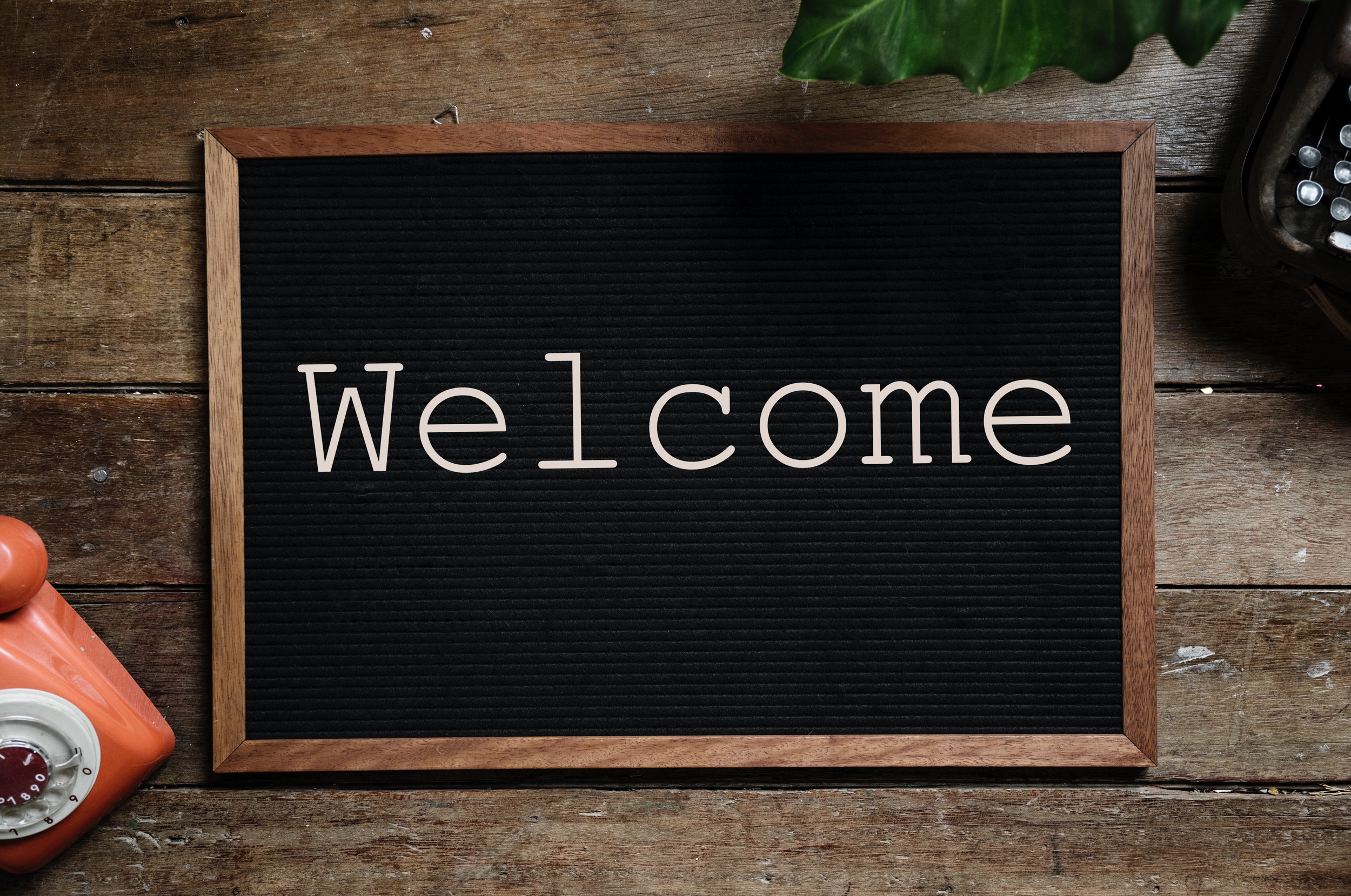 The image views a small blackboard from above on a wooden table top with the word "Welcome" written across it. There is the leaf of a plant that can be seen at the top of the image, and a coral coloured rotary style telephone in the bottom left corner, and a black one in the top right. 