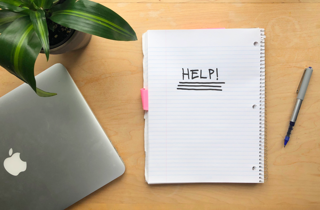 photo of a desk with a plant, laptop, pen and notebook that has "Help" written across its page 