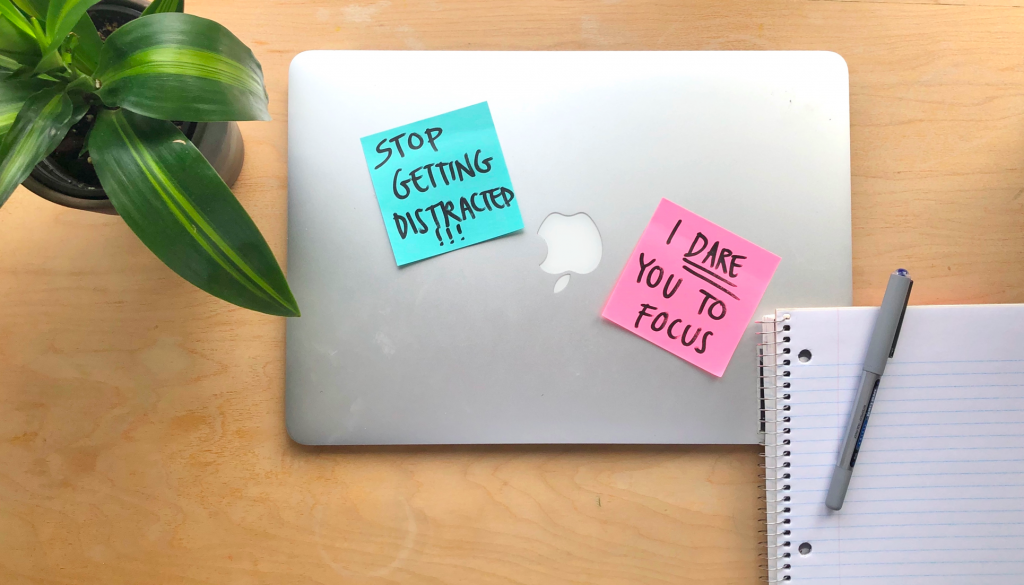 Photo of a desk with a plant, notebook, pen and laptop with sticky notes on it that read "Stop getting distracted" and "I dare you to focus."