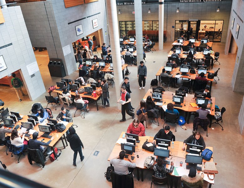 Bird's-eye view of a large open space with students sitting at communal desks and others walking through