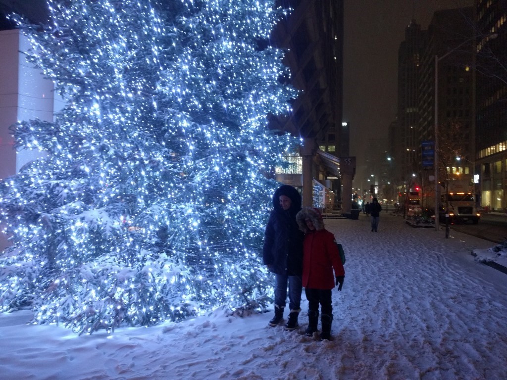 A mother and daughter standing next to a large well-lit christmas tree on a snowy, winter evening. 