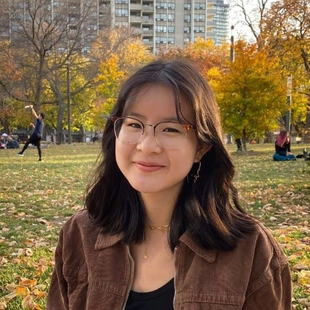 Faith is sitting outside, with trees and a cityscape in the background, wearing a dark brown jacket and black t-shirt