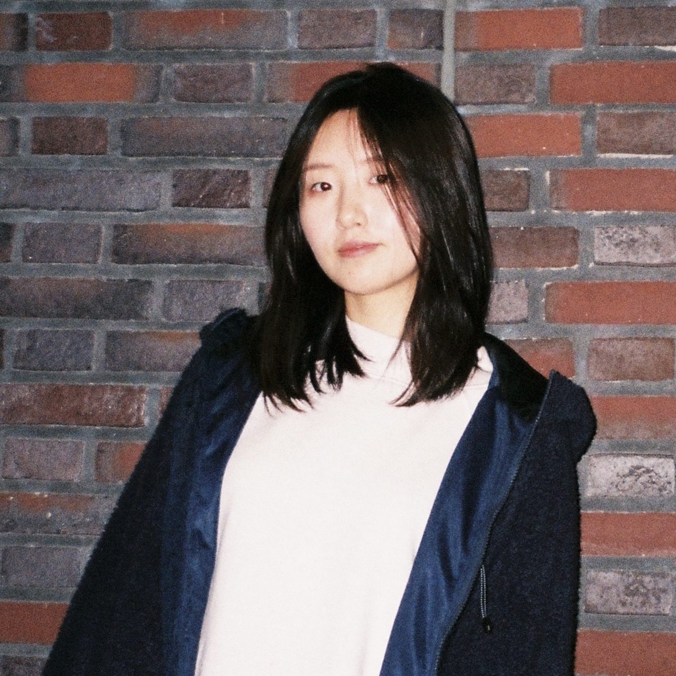 Peiyu is wearing a white shirt with a black coat, standing in front of a colourful brick wall.
