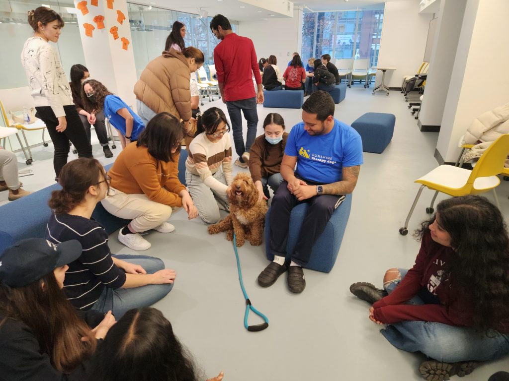 A photo of therapy dog Milo being petted by attendees with his owner on a seat beside him.