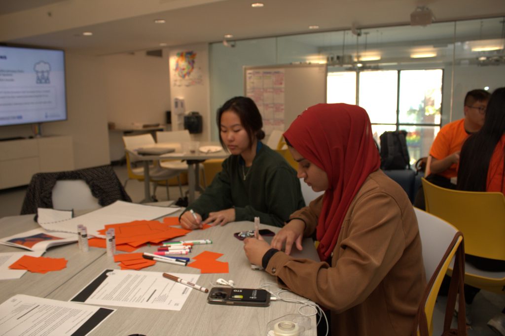 Two students sitting at a table while writing on orange pieces of paper. 