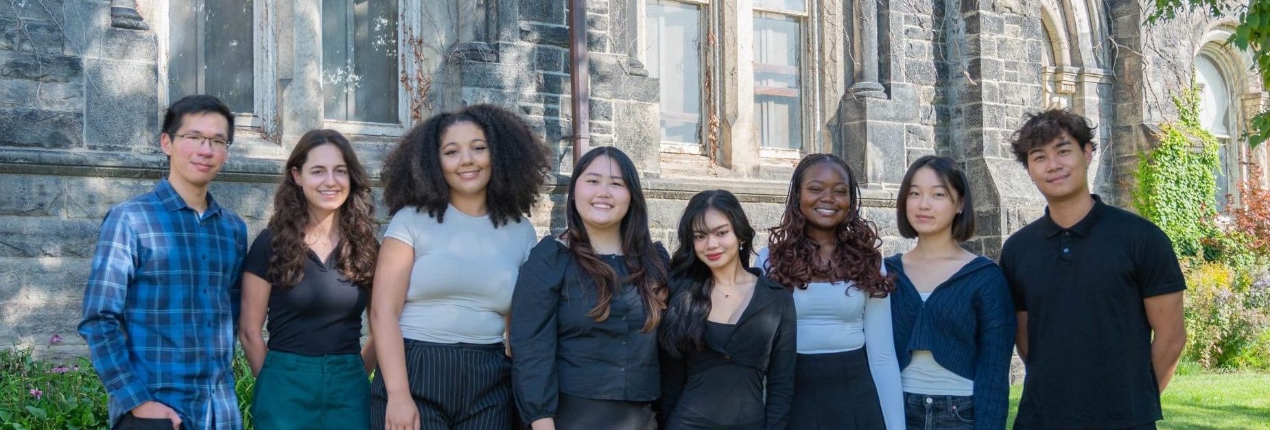 A photo of the Graphic Design team, featuring eight members standing outdoors in front of a stone building, smiling at the camera.