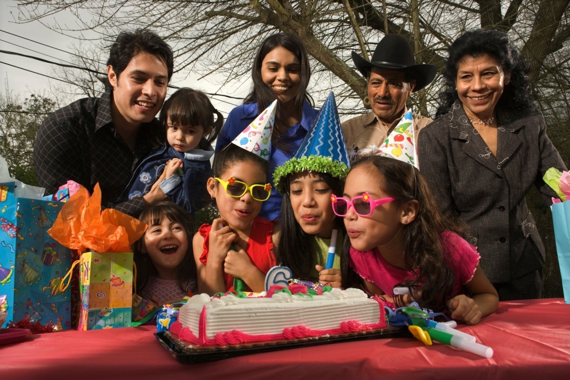 Four young children blowing out birthday candles with parents and grandparents standing behind them, in a park.