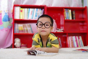 Image of child wearing large circular glasses reading a book infront of a large red bookshelf