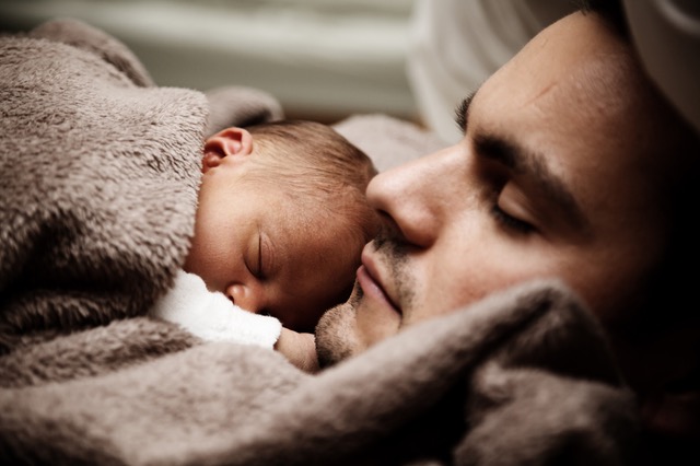 Baby sleeping on father's chest under fuzzy taupe blanket.