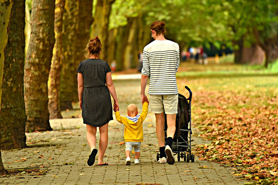 Woman and man holding baby's hand, walking with a stroller. 
