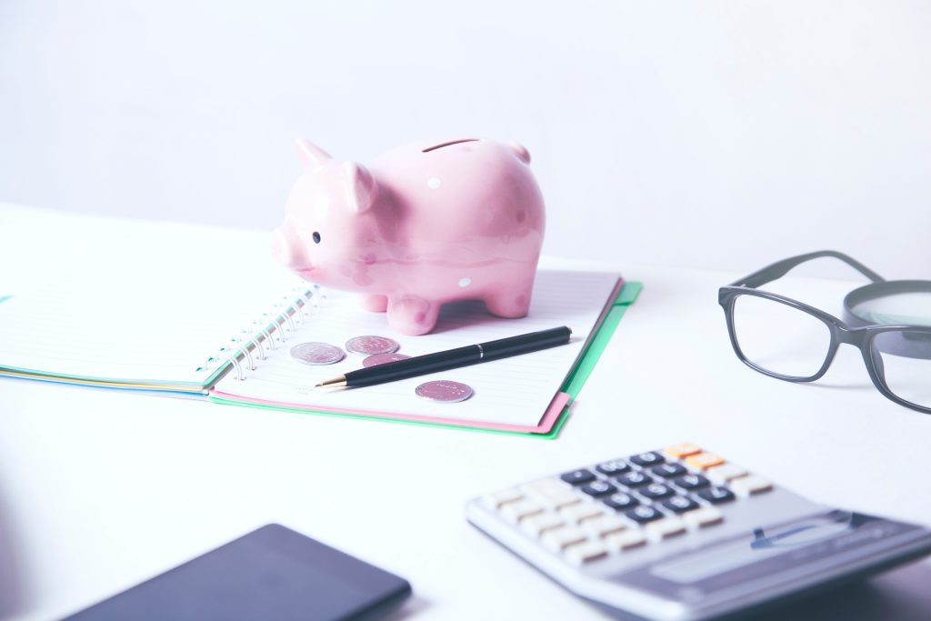 Piggy bank sitting on a notebook with a calculator, pen and glasses on the table.