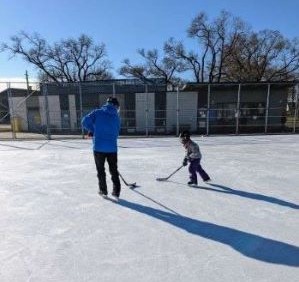 A parent and young child playing hockey at a local rink under a blue sky.