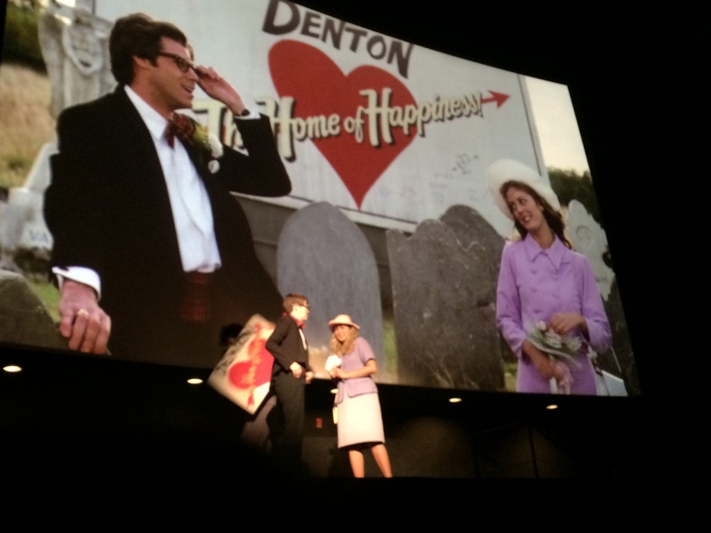 The film version of Rocky Horror Picture is playing on the screen, with Brad and Janet gazing into each others eyes. In front of the screen, the Shadow Cast for Brad and Janet are doing the same.