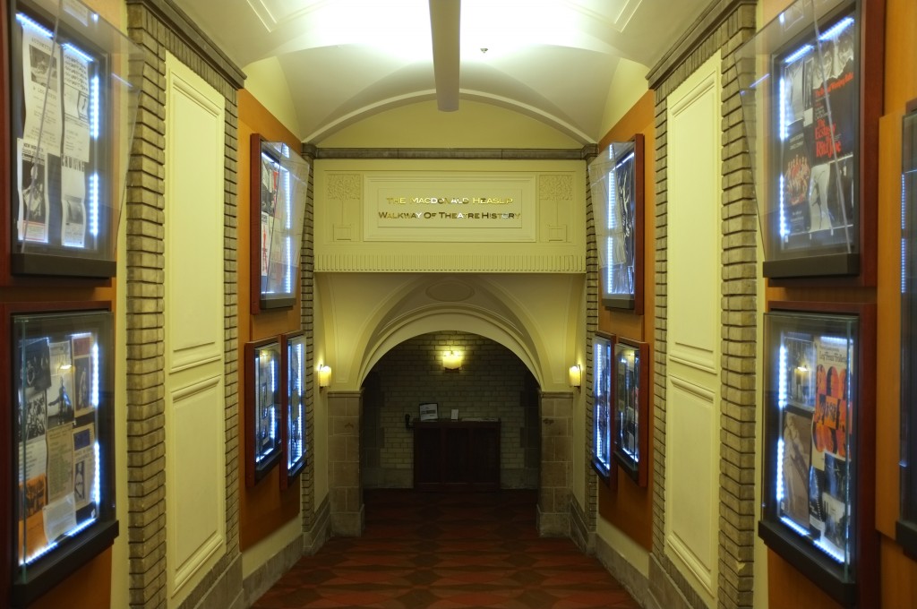 the hallway going into Hart House Theatre. the walls have shadowboxes with theatre posters. at the end of the hall is an arch. 