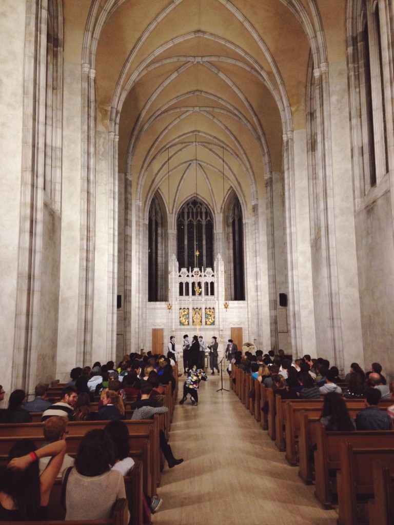 a picture of the Trinity College chapel. The pews are filled with spectators watching people dressed as mimes/clowns acting out a play at the front. 