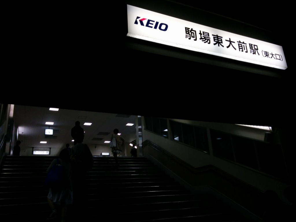 An image of the University of Tokyo, Komaba Campus Station. A bright neon sign featuring Japanese text can be seen in the top right corner of the image. Stairs leading into the station occupy the rest of the image.