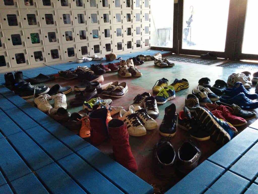 Groups of shoes haphazardly placed at the entrance to the University of Tokyo's gym. Lockers can be seen beside a door in the background.