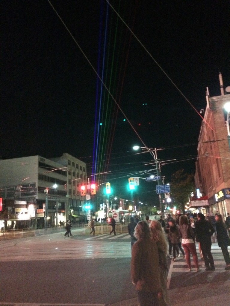 A picture of Spadina Ave. A lot of people are walking around, and there is a rainbow laser being shot across the skyline.