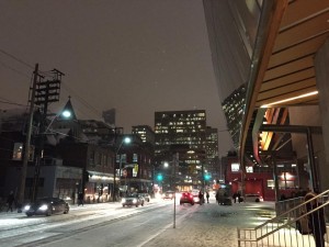 Street-level view, looking up into a snowy night sky amidst the glowing lights of towers and old Victorian houses