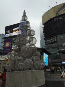 A snowy afternoon sky, surrounded by the massive bright signs on all the buildings at Yonge-Dundas Square, with a large silvery ornamental evergreen tree right in the middle