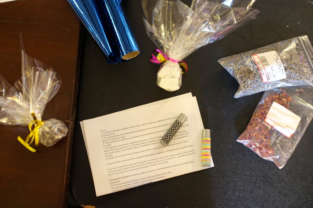 two tubes of lipbalm lie on a piece of paper that has instructions on how to make them. tubes of coloured cellophane (red and blue), two bags of scented herbs (rose and lavender) and two bath bombs wrapped in clear cellophane sit beside them. 