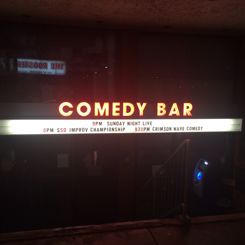 The front of the Comedy Bar. The Comedy Bar sign is glowing red in the night, and Crimson Wave is performing at 9:30pm!