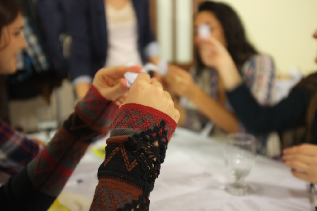 two hands in focus holding white oragami paper in the process of making a dove. blurred in the background are three more people making oragami doves. 