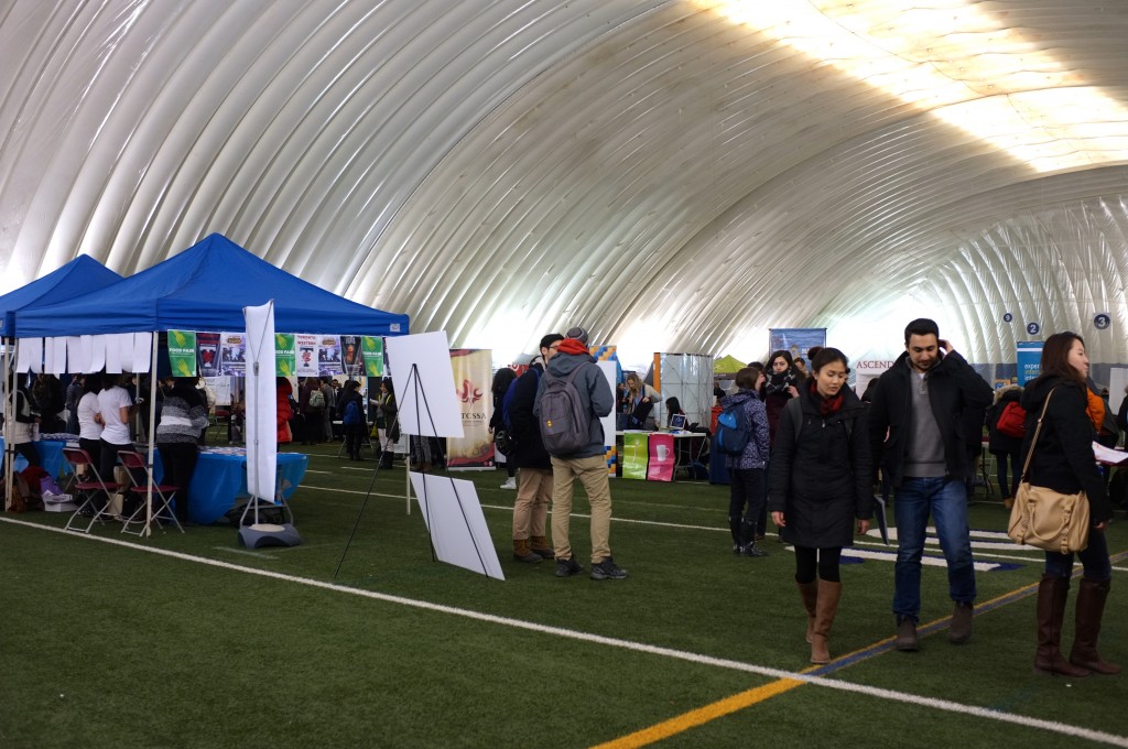 view from the entrance of the varsity dome, a large blue tent is off to the side and tables and booths can be seen in the background. Lots of people are walking around. 