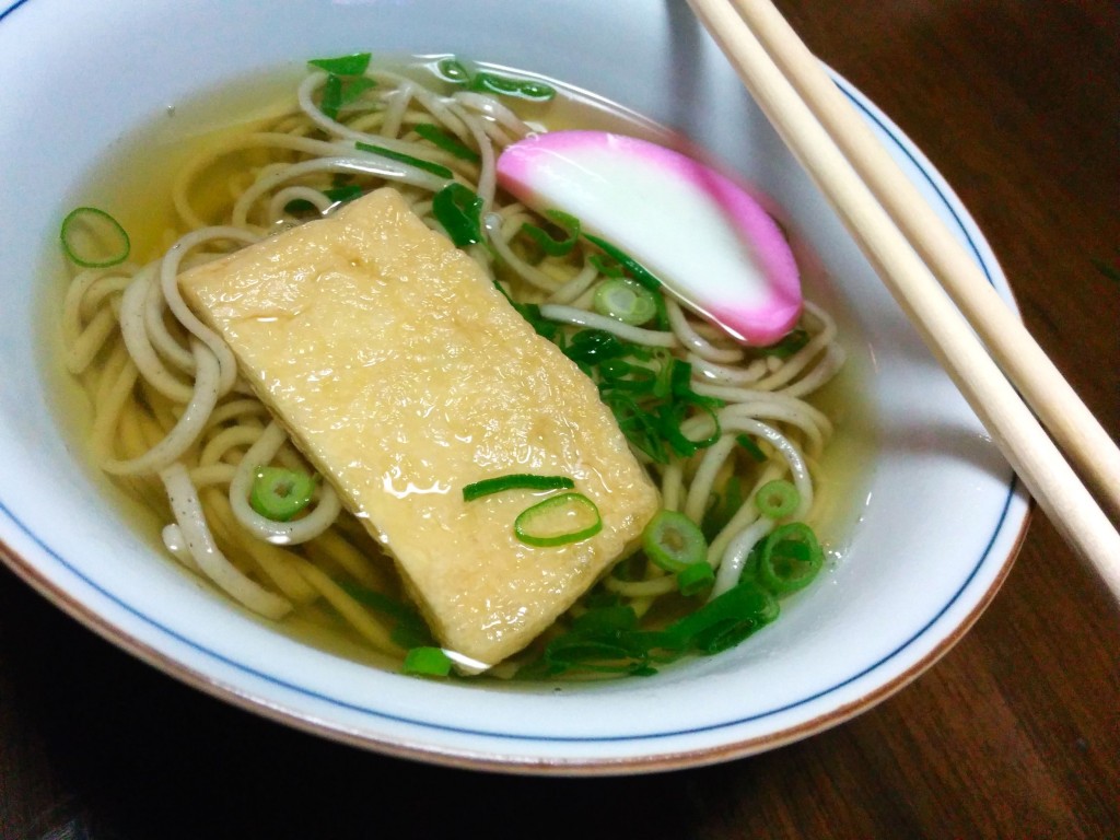 This image shows toshi-koshi soba. A slice of fish cake, tofu skin, and soba noodles are in a dark broth.