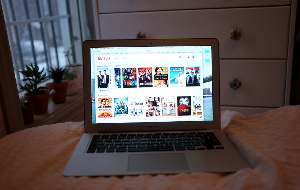a computer on a white bedspread. On the computer screen is the front page of netflix that shows suggestions to watch. 