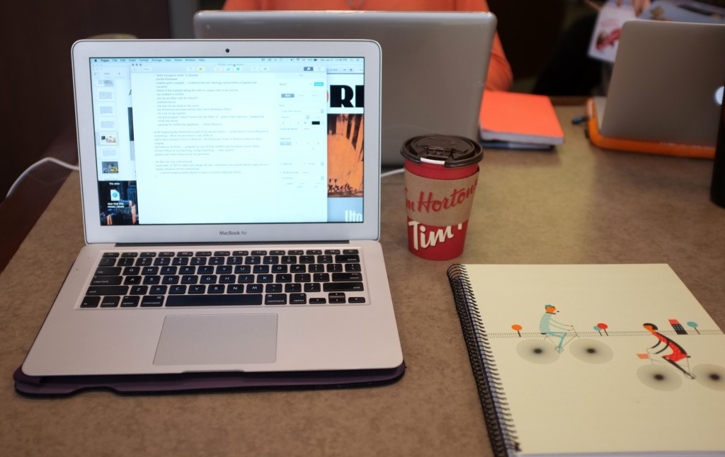 a laptop, tim hortons to go cup an a light green notebook with a drawing of two people riding bikes on it are shown on a brown table. 