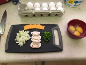 A cutting board, chopped onions and green peppers, mushrooms, and cheese slices, next to a bowl of eggs ready for mixing