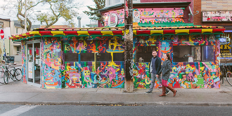 Two people walking through Kensington market , passing by a brightly painted building) 