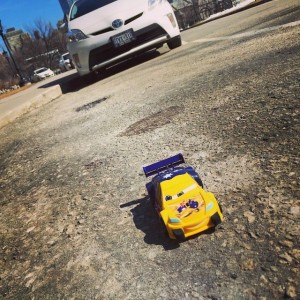 A tiny yellow toy race car with a goofy face parked in front of a real car on the road in front of University College