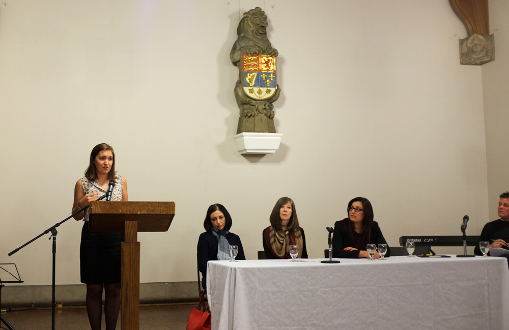 a woman stands at a podium speaking while three other women look on from their positions behind a table