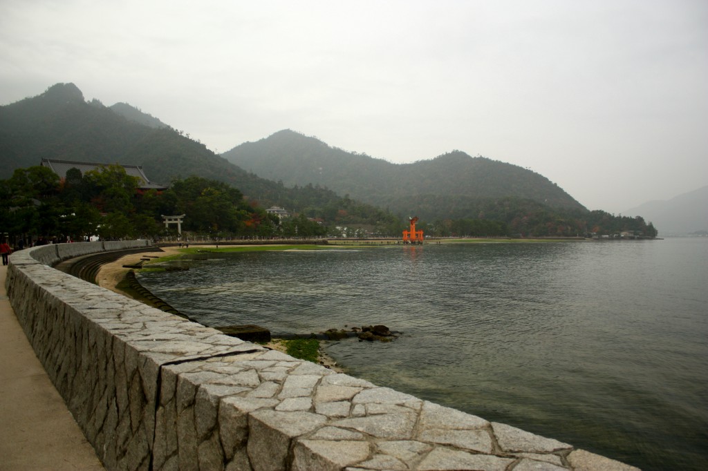 This image shows a seaside path on Miyajima island. A red toori gate is visible in the backgorund in front of a range of forested mountains.