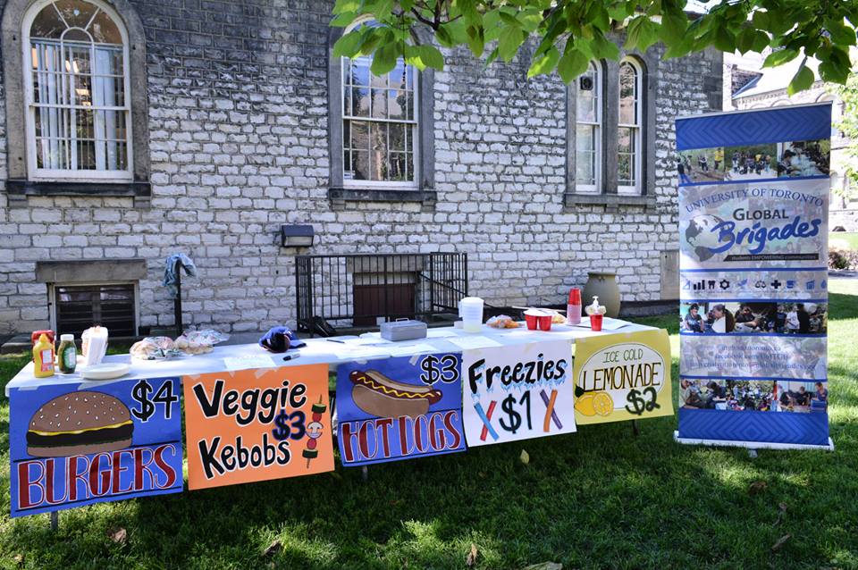 A picture of our little spread out on the UTSU lawn. Long tables with hand-painted signs advertising for burgers, hot dogs, freezies, and lemonade. Our group banner stands beside the tables.