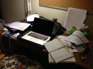 My wooden desk, covered with loose sheets and a laptop.