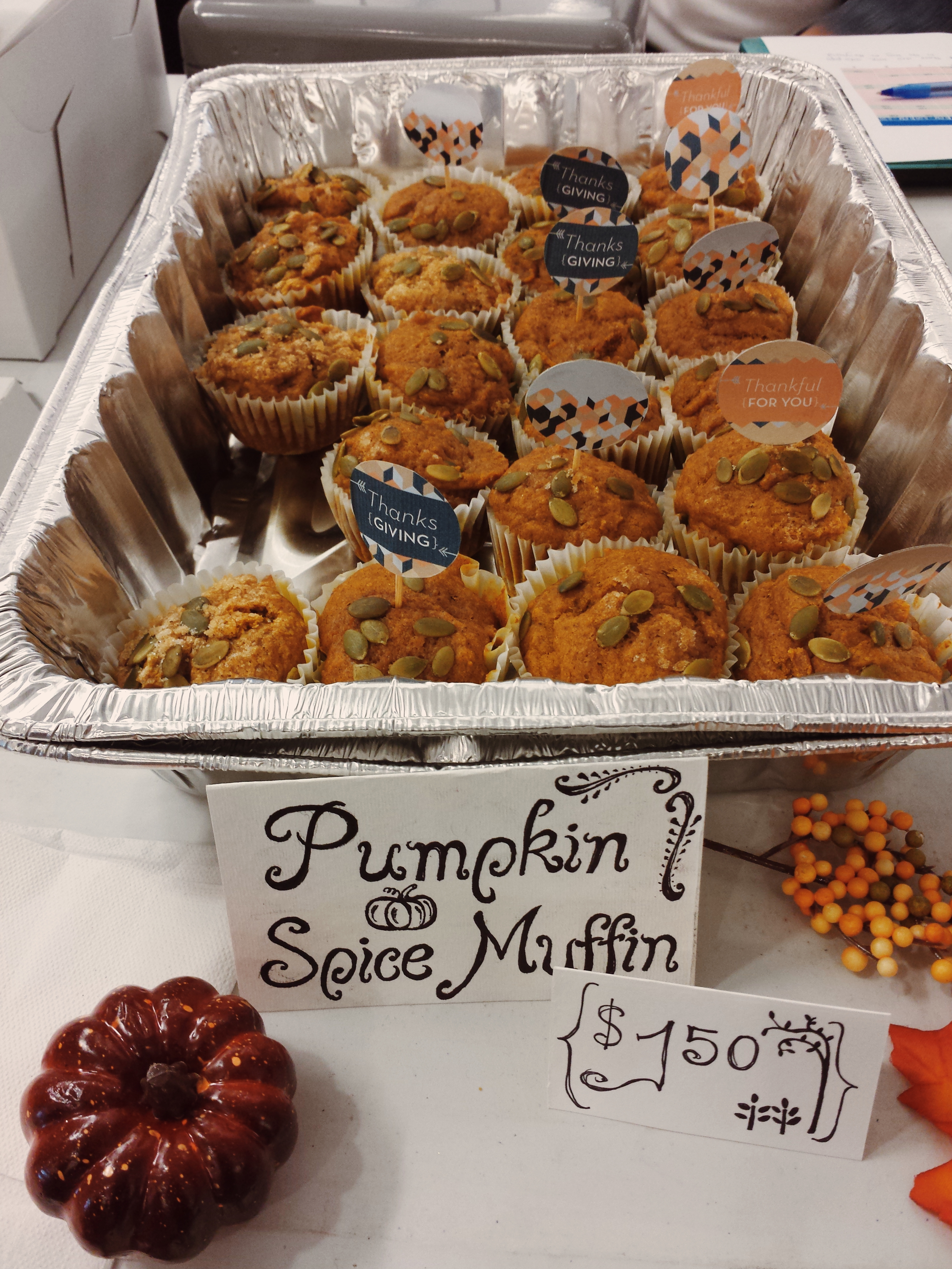 A tray of pumpkin spice muffins on a table. There is a hand-drawn sign with the price sits in front of the tray along with plastic pumpkins or squash.
