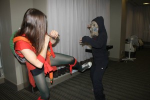 Girl in ninja turtle costume feigning a kick at someone dressed up in all black with a gas a mask -also in fighting stance.