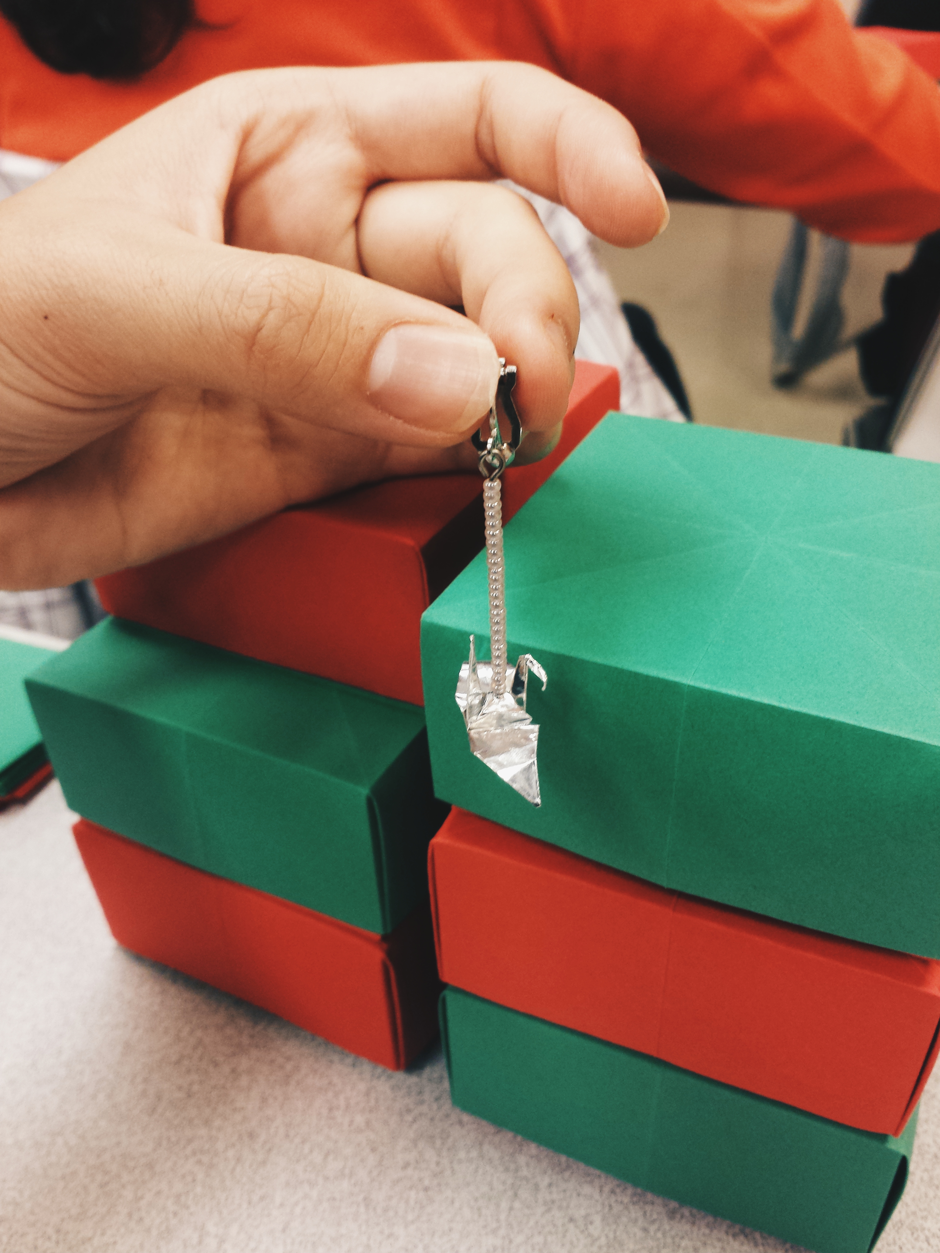 My hand holding an earring made from a small silver crane attached to a long chain earring. Red and green folded Christmas gift boxes are in the background.