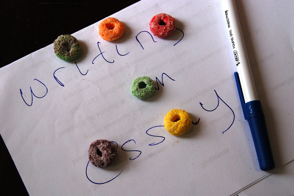 A paper with the words "writing an essay" written on it with fruit loops spread out over the text to do the "i"s and make round letter shapes.