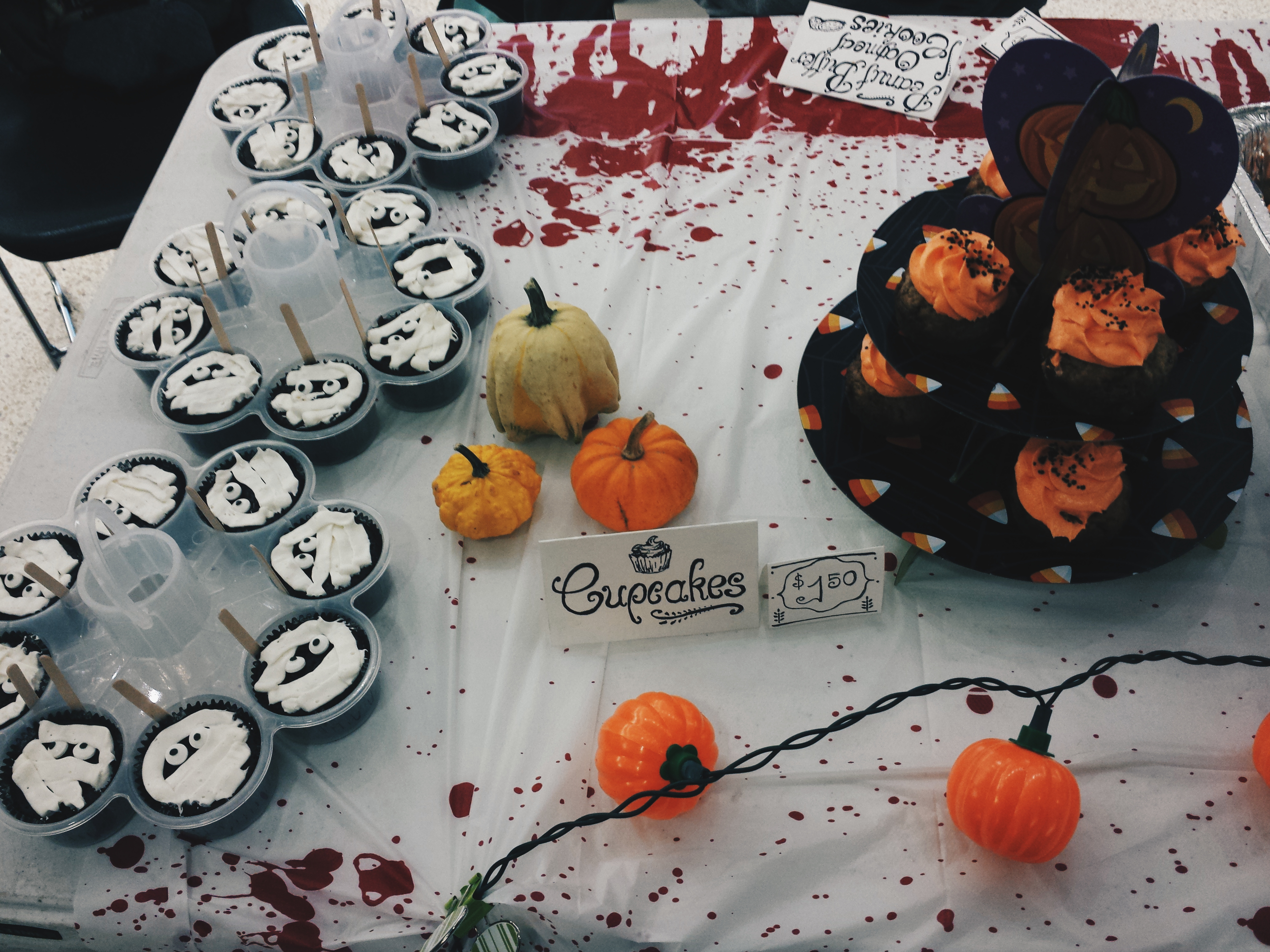 A table for a Halloween-themed bake sale, complete with a string of pumpkin lights, a blood-spattered tablecloth, and chocolate muffins decorated with white icing to look like mummies!