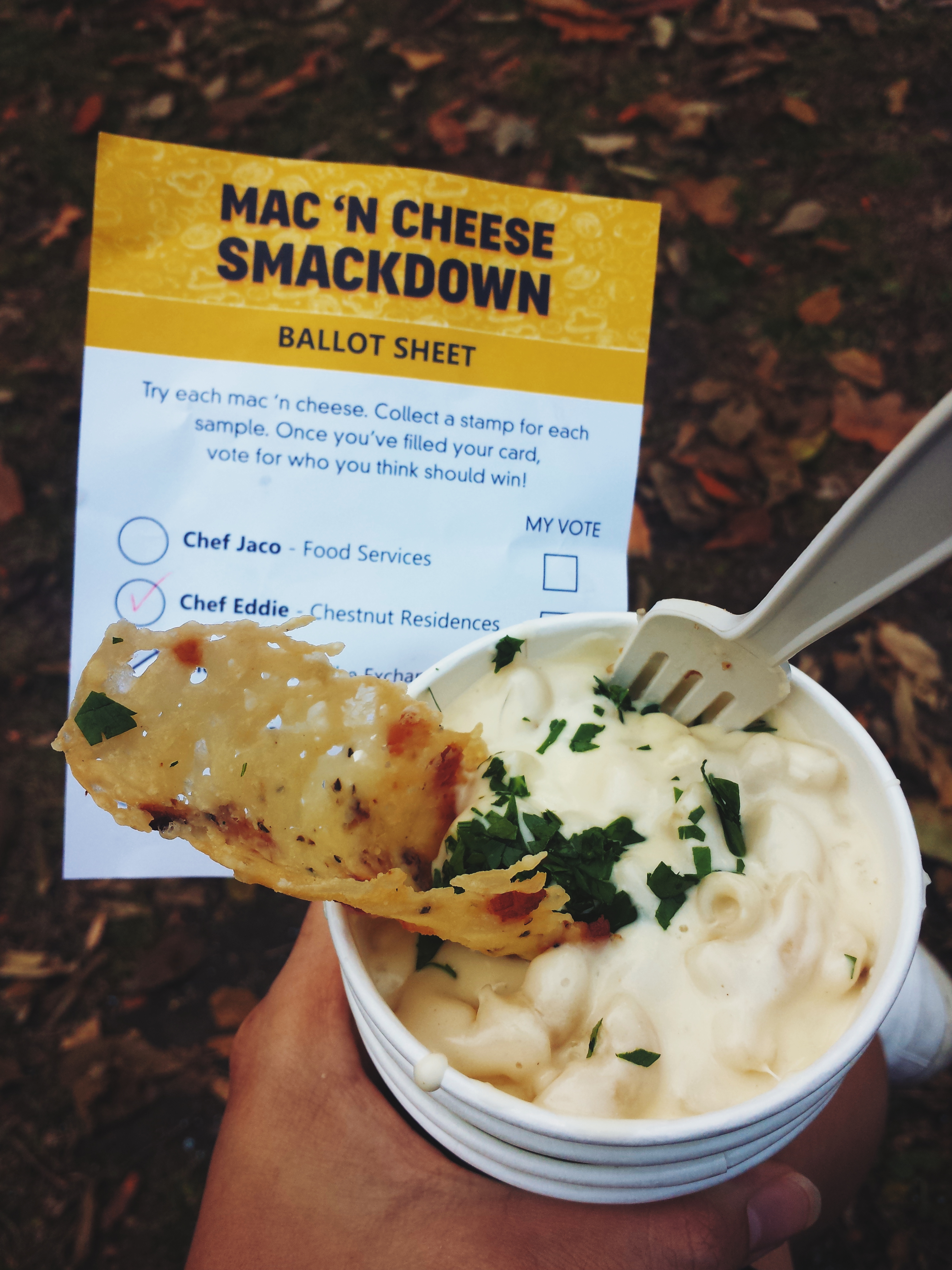 Me holding a paper bowl filled with mac and cheese and a fried cheese fritter on top. In the background is the ballot sheet with all the names of the chefs who participated in the event.