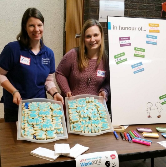 A pop-up booth with two Student Life staffers holding up trays of M-shaped cookies, next to sign that reads "in honor of..." with stickers with the names of mentors.