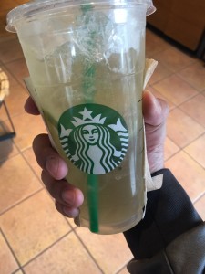 a photo of a hand holding a yellow coloured iced beverage in a plastic cup with a green straw and a logo with the image of a white mermaid in the middle of a green circle