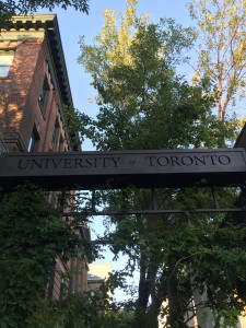 A photo a bar with an etching that reads University of Toronto in front of a red brick building and a tree