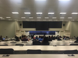 a photo of a lecture hall with tiered rows of black chairs and grey tables facing a lecture stand and long table with a three side by side blackboards and three large screens side by side above the blackboards. The photo also shows a tiled ceiling with uniform rows of fluorescent lighting and off-white walls. There are several students sitting in the seats while a professor is scribbling something on the blackboar.
